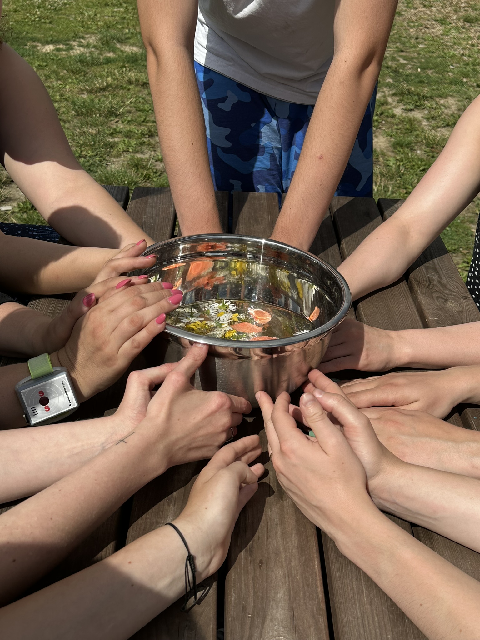 Ruce účastníků okolo kovové mísy s vodou a barevnými květy.